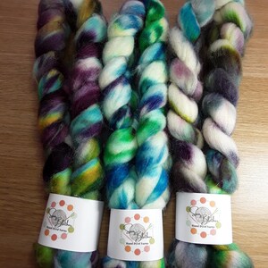 May include: Three skeins of hand-dyed yarn in vibrant colours. The yarn is twisted into thick, textured ropes, with hues of purple, green, blue, and yellow. Each skein has a white label with the text "Spin Blathers Hand Dyed Yarns".