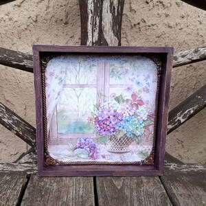 Peut inclure: Une impression encadrée d'une peinture à l'aquarelle représentant une fenêtre avec vue sur un jardin. La fenêtre est ouverte et il y a des fleurs dans un panier sur le rebord de la fenêtre. Les fleurs sont violettes, bleues et blanches.