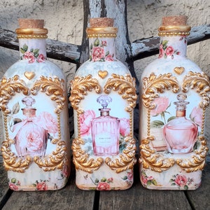 Op de afbeelding: Drie decoratieve glazen flessen met gouden accenten en bloemmotieven. Elke fles heeft een hartvormig ontwerp en een kleinere fles met een bloemmotief erin. De flessen staan op een houten oppervlak.