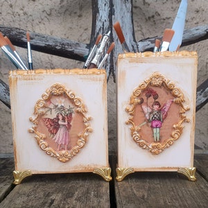 Könnte beinhalten: Zwei weiße dekorative Holzkisten mit goldenen Verzierungen und Feenillustrationen. Die Kisten sind für Stifte, Bleistifte oder andere kleine Gegenstände gedacht.