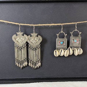 May include: Three pairs of silver earrings with different designs. The first pair features a heart-shaped pendant with intricate details and a chain of dangling beads. The second pair has a square pendant with colorful accents and a chain of dangling seashells. The third pair is similar to the first, but with a slightly different design.