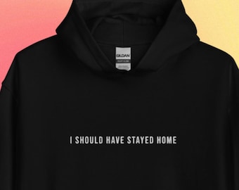 May include: Black hoodie with the white text "I SHOULD HAVE STAYED HOME". The sweatshirt features a front pocket and a drawstring hood. The background is a gradient of pink, yellow, and orange.
