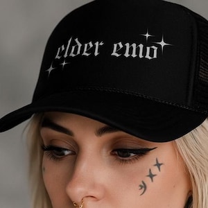 May include: Black trucker hat with the text "elder emo" in white gothic lettering. The hat has a mesh back and a curved brim. The hat is worn by a person with tattoos and piercings.
