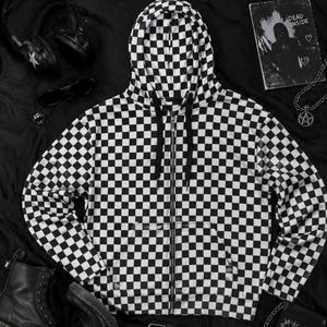 May include: A black and white checkered zip-up hoodie featuring a hood with black drawstrings, long sleeves, and a front zipper closure. The garment is covered in a repeating square pattern throughout the entire fabric, including the hood and sleeves.