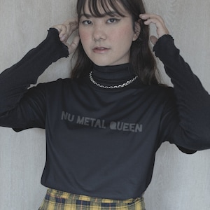 May include: A black long-sleeve shirt with the text "NU METAL QUEEN" in a dark grey font. The shirt is paired with a silver chain necklace and a yellow and black plaid skirt.