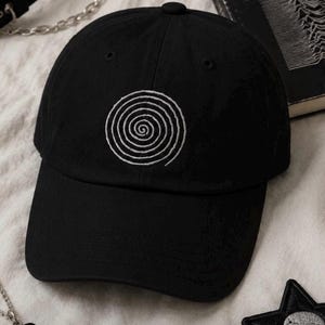 May include: A black baseball cap featuring a white embroidered spiral design on the front panel. The cap has a structured crown, a curved brim, and eyelets for ventilation. The design is centered on the front of the cap, providing a clear visual focal point.