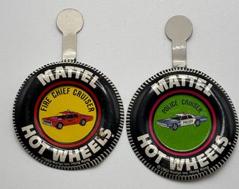 Può includere: Due badge di auto giocattolo Hot Wheels in bianco e nero con un cruiser di capo dei pompieri rosso e giallo e un cruiser di polizia blu e verde. I badge sono rotondi e hanno un fermaglio in metallo argentato nella parte superiore. Il testo sui badge recita "Mattel Fire Chief Cruiser Hot Wheels ©1969 Mattel Inc. Hong Kong" e "Mattel Police Cruiser Hot Wheels ©1968 Mattel Inc. Hong Kong".