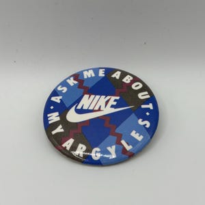 May include: A blue and white button with the Nike logo and the text "Ask Me About Argyle's" around the edge.