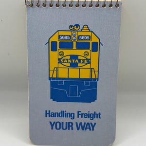May include: A spiral-bound notepad with a blue cover featuring a yellow and blue illustration of a Santa Fe train engine. The notepad has the text "Handling Freight YOUR WAY" in blue.