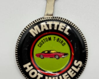 May include: A black and white Hot Wheels button with a red and green circle in the center. The circle features a red car with a yellow interior and the text "Custom T-Bird". The button has a silver metal loop on top.