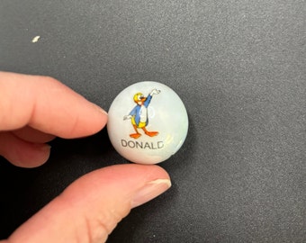 May include: A white marble featuring a cartoon image of Donald Duck in a blue jacket, yellow hat, and orange feet. The word "DONALD" is printed below the character. The marble is held by a hand.