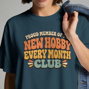 May include: A dark teal crew neck t-shirt featuring the text "PROUD MEMBER OF THE NEW HOBBY EVERY MONTH CLUB" in a multi-colored, wavy, retro-style font. The lettering uses shades of orange, yellow, and light green against the dark fabric.