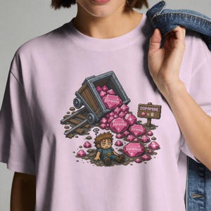 Puede incluir: Una camiseta de color morado claro con un gráfico de pixel art de una vagoneta de mina volcada que derrama cristales rosas etiquetados como 'DOPAMINE'. Un personaje cansado está sentado en el suelo junto a un letrero de madera que dice 'DOPAMINE x 0'.