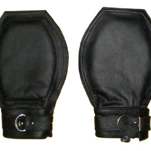 May include: A pair of black leather restraints. Each restraint has a padded, hexagonal top and a wrist strap with a metal buckle. The leather appears smooth and the stitching is visible around the edges.