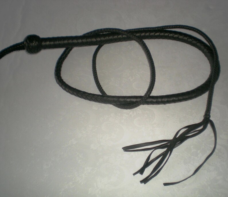 May include: A black leather bullwhip with a braided handle and a long tassel. The whip is coiled and laid out on a white surface.