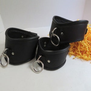 May include: Three black leather collars with silver metal rings. The collars are on a white surface with yellow shredded paper in the background.