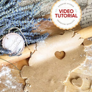 May include: A close-up of a brown gingerbread cookie dough rolled out on a wooden surface. The dough has a heart-shaped cutout and is surrounded by white powdered sugar. A wooden rolling pin and a small heart-shaped cookie are also visible in the image.