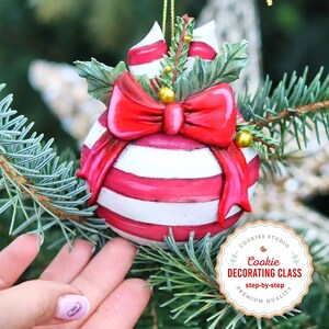 May include: A red and white striped Christmas ornament with a red bow and green holly. The ornament is hanging on a Christmas tree. The text on the ornament reads "Cookie Decorating Class step-by-step Premium Quality".