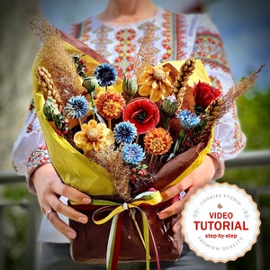 May include: A vibrant bouquet of handmade flowers, featuring red poppies, blue and yellow dahlias, and wheat, wrapped in yellow paper and tied with a ribbon. The bouquet is held by a person wearing a white embroidered shirt. The image includes a logo with the text "VIDEO TUTORIAL".