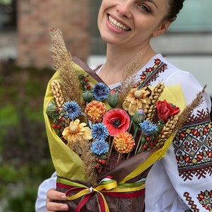 May include: A vibrant bouquet of handmade flowers, featuring red poppies, blue cornflowers, and orange dahlias, wrapped in brown paper and tied with ribbons. The bouquet includes wheat stalks and dried grasses. The person holding the bouquet is wearing a white embroidered shirt.