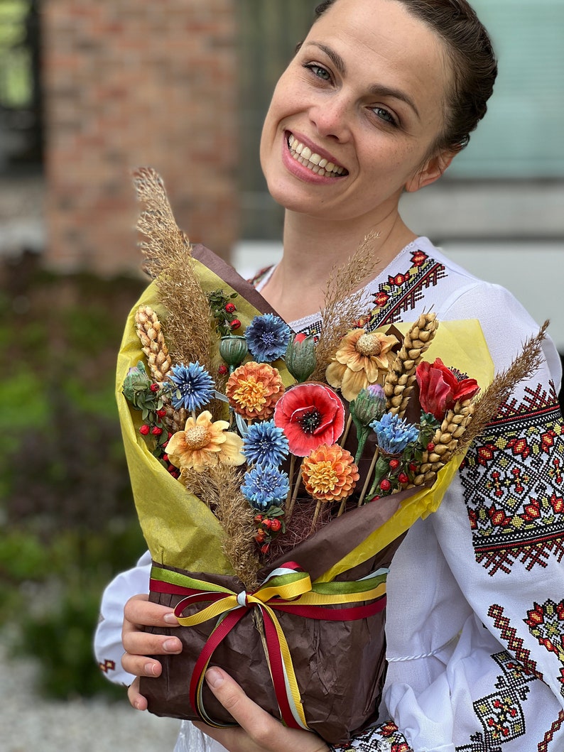 May include: A vibrant bouquet of handmade flowers, featuring red poppies, blue cornflowers, and orange dahlias, wrapped in brown paper and tied with ribbons. The bouquet includes wheat stalks and dried grasses. The person holding the bouquet is wearing a white embroidered shirt.