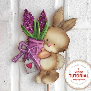 May include: A brown bunny holding a purple vase with a pink bow and a heart-shaped tag. The vase contains a bouquet of purple hyacinths. The bunny is standing on a white brick background. The image includes the text "COOKIES STUDIO VIDEO TUTORIAL step-by-step PREMIUM QUALITY".