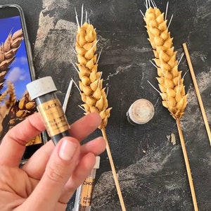 May include: Image of wheat stalks, possibly for cake decoration, with a golden colour. A phone displays a wheat field image. Small bottles of edible food colouring are also visible, including a bottle labelled "BLACK".