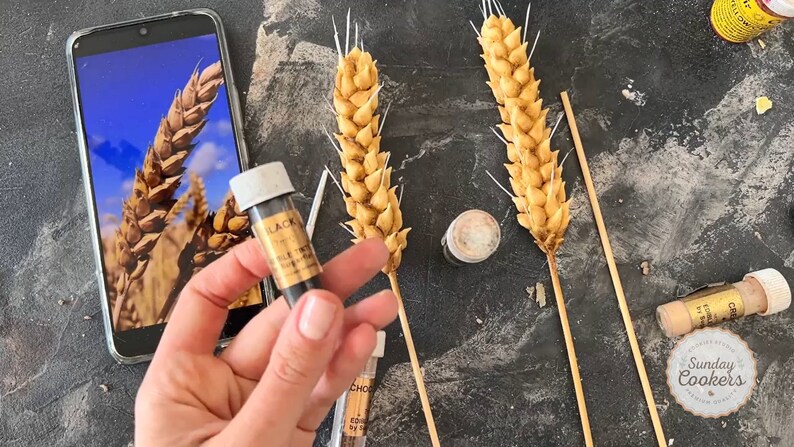 May include: Image of wheat stalks, possibly for cake decoration, with a golden colour. A phone displays a wheat field image. Small bottles of edible food colouring are also visible, including a bottle labelled "BLACK".