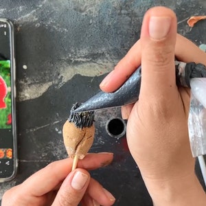 May include: A person is decorating a poppy-shaped cake with black icing from a piping bag. A smartphone displays a photo of a red poppy flower. The image also shows a finished poppy and the text "SUNDAY COOKERS".