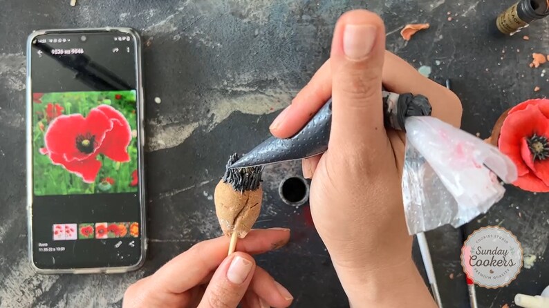 May include: A person is decorating a poppy-shaped cake with black icing from a piping bag. A smartphone displays a photo of a red poppy flower. The image also shows a finished poppy and the text "SUNDAY COOKERS".