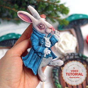 Könnte beinhalten: Ein weißer Hasen-Lebkuchen, verziert mit blauem Zuckerguss und einem weißen Kragen. Der Lebkuchen hält eine Taschenuhr in seiner rechten Hand. Der Lebkuchen befindet sich auf einem weißen Hintergrund mit einem grünen Pilz und einer weißen Rose im Hintergrund. Der Text "VIDEO TUTORIAL step-by-step PREMIUM QUALITY" befindet sich auf einem braunen Kreis in der unteren rechten Ecke des Bildes.
