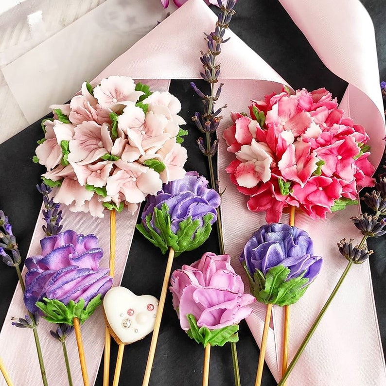 May include: Assortment of colorful faux flower arrangements on sticks. The flowers are pink, purple, and white. The flowers are arranged in a bouquet and are on a white and pink background.