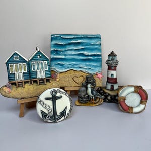 May include: A set of five decorative cookies with a nautical theme. The cookies are painted to look like a beach scene, a lighthouse, a life preserver, an anchor, and two beach houses.