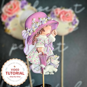 May include: A purple and white decorated gingerbread cookie of a girl wearing a pink hat and holding an umbrella. The girl is standing on a stick and has a white kitten beside her. The cookie is decorated with flowers and has a floral pattern on the dress. The cookie is surrounded by a white background with the text "Cookies Studio Video Tutorial step-by-step Premium Quality" in a circle.