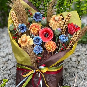 May include: A vibrant bouquet of artificial flowers, featuring red poppies, blue cornflowers, and orange dahlias, combined with wheat and wrapped in brown and yellow paper. The bouquet is tied with a multi-coloured ribbon.