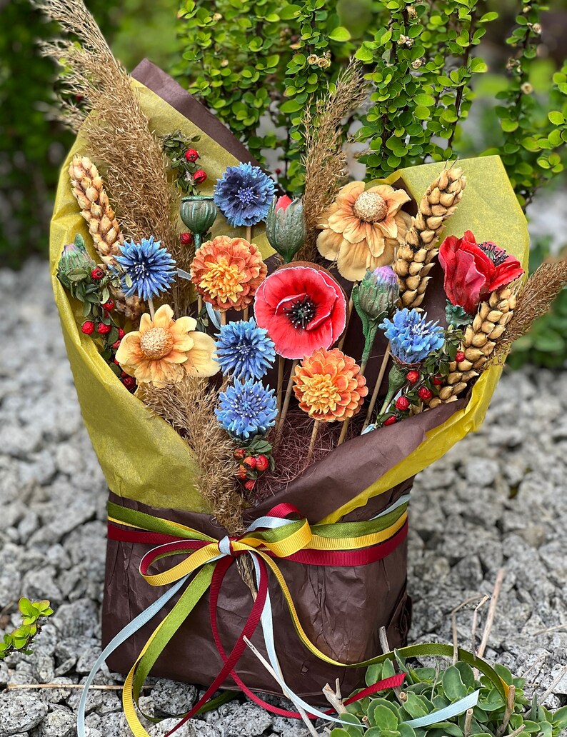 May include: A vibrant bouquet of artificial flowers, featuring red poppies, blue cornflowers, and orange dahlias, combined with wheat and wrapped in brown and yellow paper. The bouquet is tied with a multi-coloured ribbon.