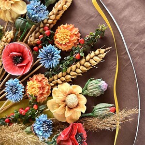 May include: A vibrant floral display with handmade flowers in red, orange, yellow, and blue. The bouquet includes poppies, dahlias, and other blooms, with wheat and berries, arranged on a brown surface. A decorative piece with a natural theme.