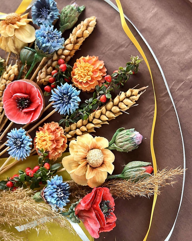 May include: A vibrant floral display with handmade flowers in red, orange, yellow, and blue. The bouquet includes poppies, dahlias, and other blooms, with wheat and berries, arranged on a brown surface. A decorative piece with a natural theme.