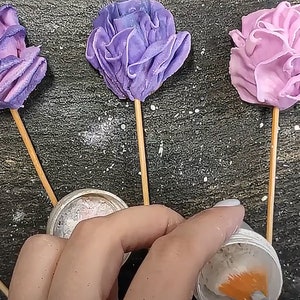 May include: Four purple and pink buttercream flowers on sticks, with a container of edible glitter and a container of orange food coloring.