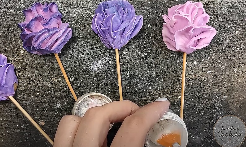 May include: Four purple and pink buttercream flowers on sticks, with a container of edible glitter and a container of orange food coloring.