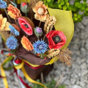 May include: A vibrant bouquet of artificial flowers, featuring red poppies, blue cornflowers, and orange daisies. The flowers are wrapped in brown paper with a yellow accent and tied with ribbons. The arrangement is set against a backdrop of green foliage.