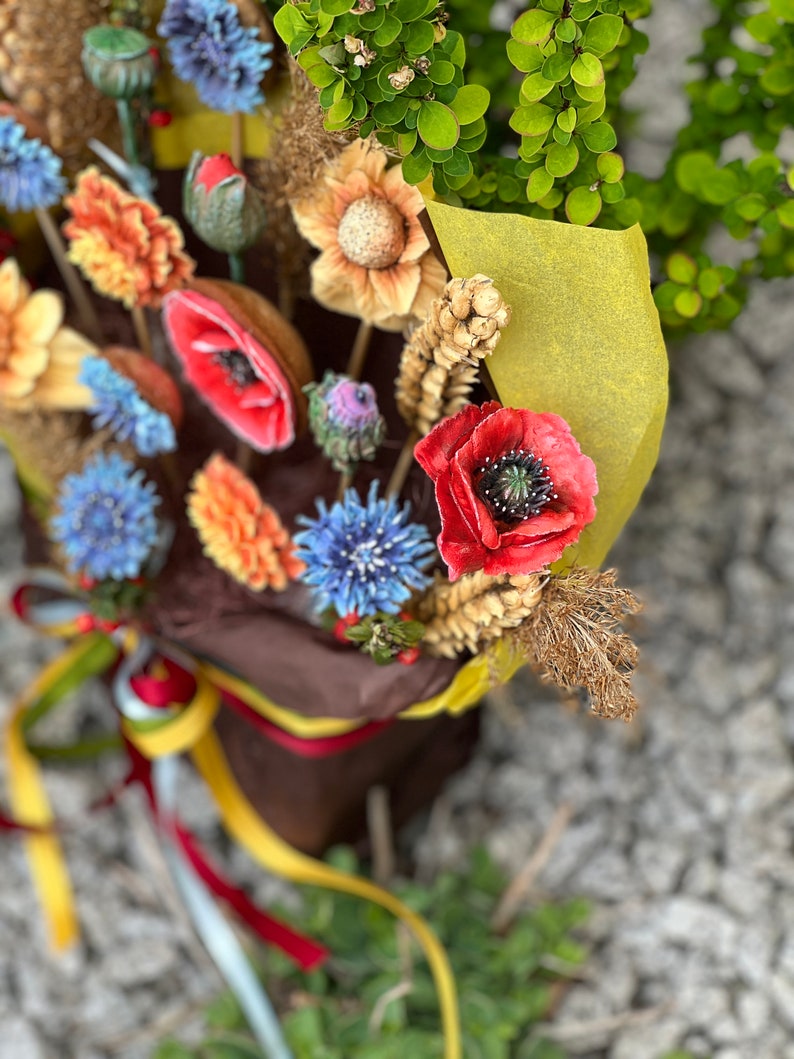 May include: A vibrant bouquet of artificial flowers, featuring red poppies, blue cornflowers, and orange daisies. The flowers are wrapped in brown paper with a yellow accent and tied with ribbons. The arrangement is set against a backdrop of green foliage.