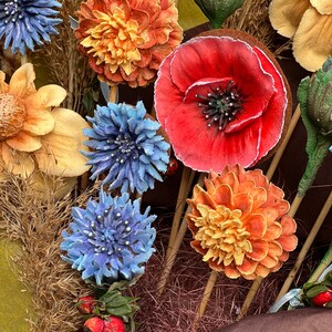 May include: Close-up of a collection of artificial flowers in various colours. The arrangement includes orange, red, and blue flowers, with some yellow and green accents. The flowers are on wooden sticks, creating a bouquet.