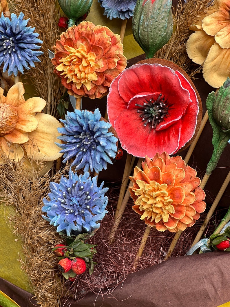 May include: Close-up of a collection of artificial flowers in various colours. The arrangement includes orange, red, and blue flowers, with some yellow and green accents. The flowers are on wooden sticks, creating a bouquet.