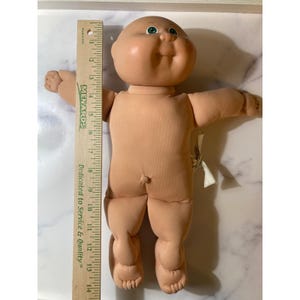 Könnte beinhalten: Eine Vintage Cabbage Patch Kid Puppe mit einem pfirsichfarbenen Vinylkopf und einem weichen Körper. Die Puppe hat grüne Augen und einen lächelnden Ausdruck. Ein Holzlineal wird zur Messung neben die Puppe gelegt. Die Puppe ist etwa 35,5 cm groß.