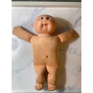 May include: A vintage Cabbage Patch Kid doll with a peach-colored face and body. The doll has dark eyes, a smiling expression, and outstretched arms. The doll's body is made of soft fabric.