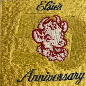 May include: A yellow embroidered patch featuring a white cartoon cow wearing a red flower lei. The patch reads "Elsie's 50th Anniversary" in blue embroidery.