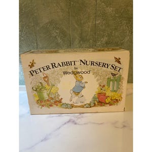 Puede incluir: Una caja vintage del juego Peter Rabbit Nursery Set de Wedgwood. La caja presenta ilustraciones de Peter Rabbit, herramientas de jardinería y flores. La caja es de color crema con el texto "Peter Rabbit Nursery Set by Wedgwood".