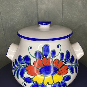 Puede incluir: Cazuela de cerámica blanca con un diseño floral azul y blanco. La cazuela tiene una tapa y dos asas a los lados. El diseño floral presenta flores rojas, amarillas y azules.