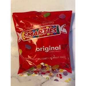 Op de afbeelding: Een rode zak met Smarties originele snoeprollen. De zak heeft het Smarties-logo, de woorden "original candy rolls" en de tekst "Family Owned Since 1949". De zak vermeldt ook dat de snoep glutenvrij en pindavrij is.